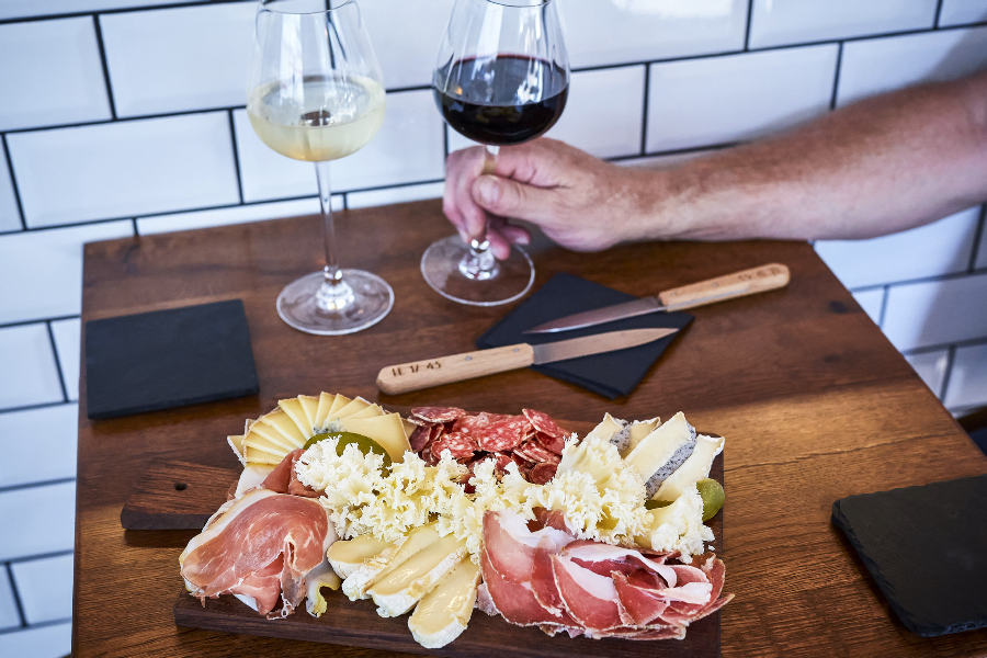 Personne assise à table tenant un verre de vin, devant une planche mixte de charcuterie et fromage - ©Spassoff